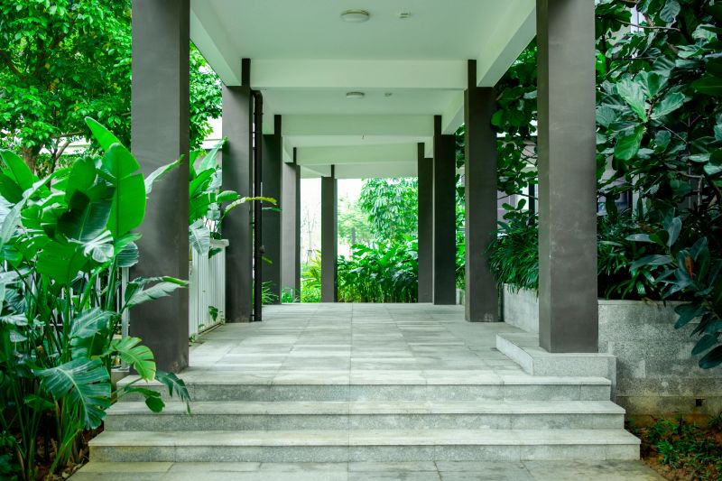A modern covered walkway with dark concrete columns and stone steps, surrounded by lush tropical greenery including large-leaf plants and trees. The scene blends minimalist architecture with abundant nature, creating a peaceful, refreshing urban oasis.