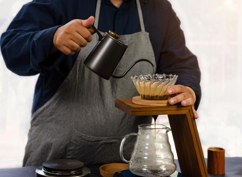 Barista in navy shirt and gray apron manual drip coffee brewing by pouring hot water from black gooseneck kettle through glass dripper into clear glass carafe below in cafe