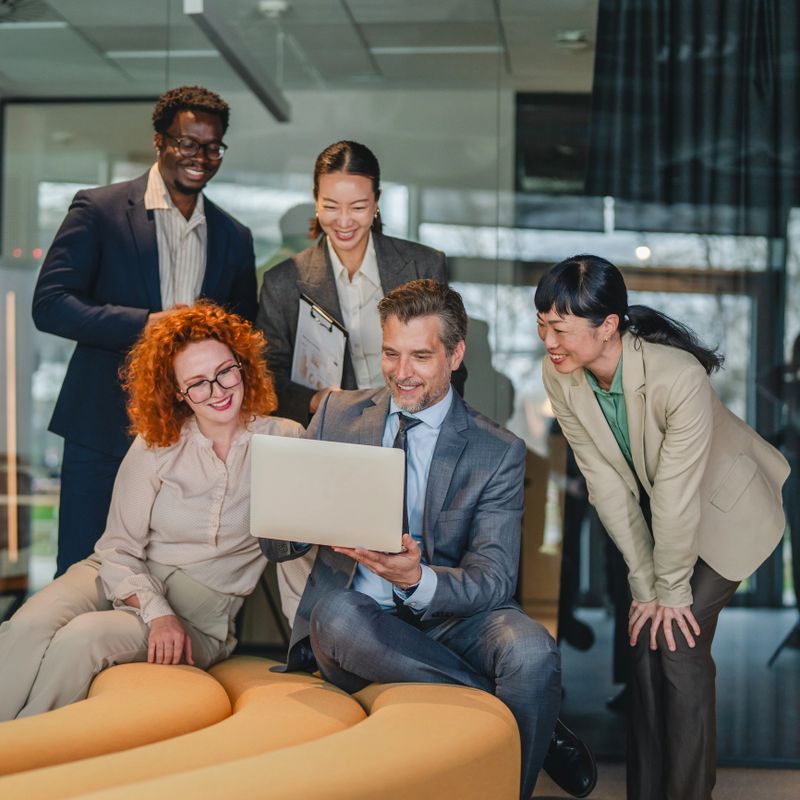 Diverse business professionals gathering around a man and a woman looking at laptop screen, collaborating and discussing ideas together in modern office space
