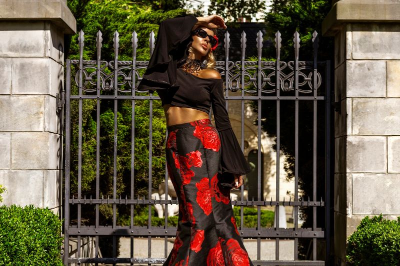 Romantic woman in elegant black and red floral gown standing outside in front of a metal gate in a palace garden on a sunny day