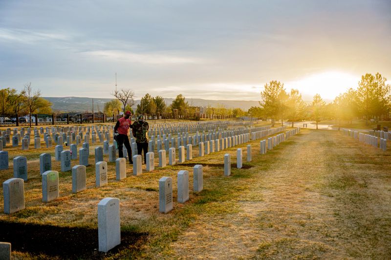 Two elderly African American male veterans stand among rows of grave markers at a national cemetery, dwarfed by a dramatic golden sunset flooding the frame with light. Headstones stretch to the horizon with trees and mountains in the distance under a glowing evening sky.