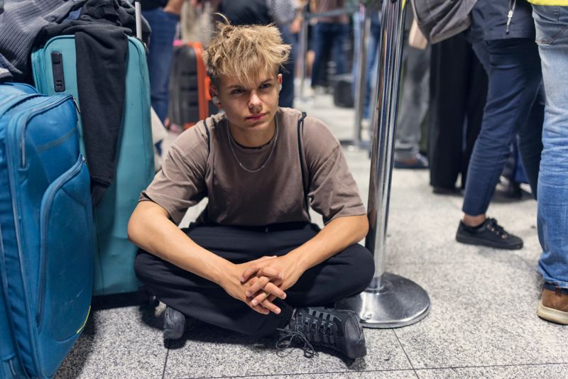 A compelling, candid shot of a young male traveler sitting on the terminal floor while waiting in a long queue. Surrounded by large suitcases and the legs of other passengers, he sits cross-legged with a stoic expression, wearing wireless earbuds to tune out the airport noise. The low-angle perspective emphasizes the feeling of exhaustion and the reality of long layovers, flight delays, or slow security checks. The lighting is natural and atmospheric, capturing a relatable moment for young travelers in a busy, modern transit environment.Shot with Canon R5