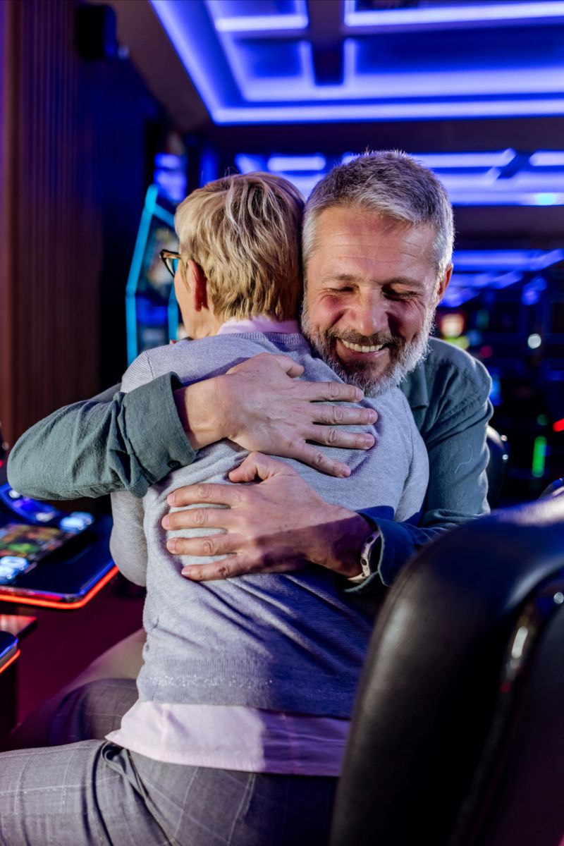A joyful senior couple shares a celebration hug in a vibrant modern casino with glowing lights. The emotional connection highlights joy and accomplishment within the dynamic gaming atmosphere.