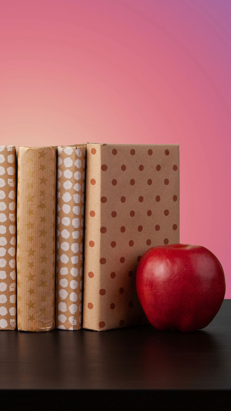 Four books wrapped in paper sit on a table next to a red apple and a colorful background.