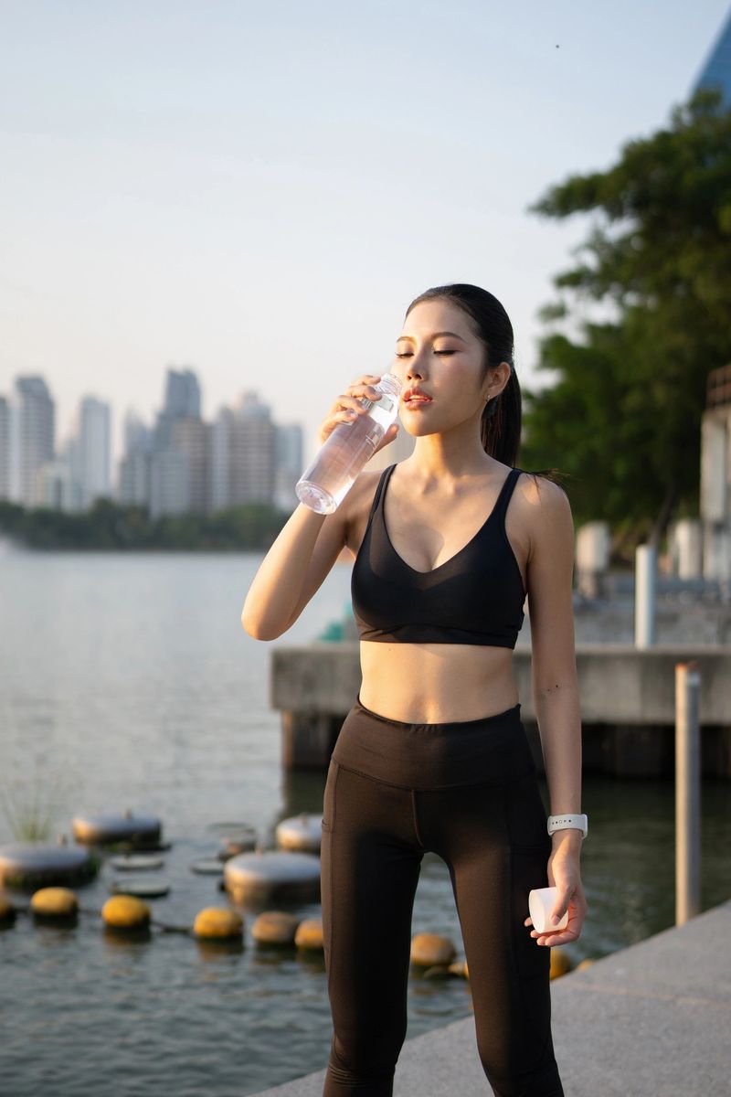 Happy asian woman wearing sportswear, drinking a water bottle, smiling after a working out in an urban city environment on a sunny day, promoting a healthy lifestyle and hydration.