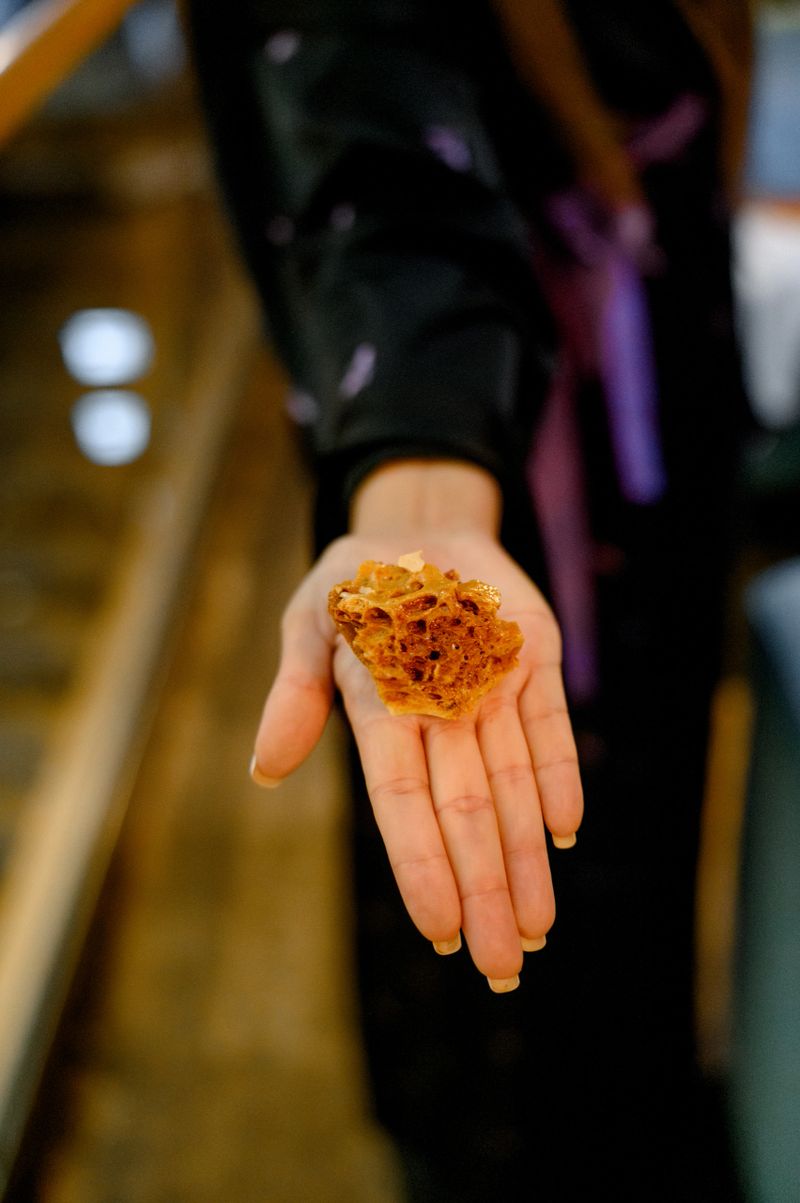 A hand displays a piece of amber gemstone, showcasing its rich color and texture, with a blurred background of natural elements and soft lighting