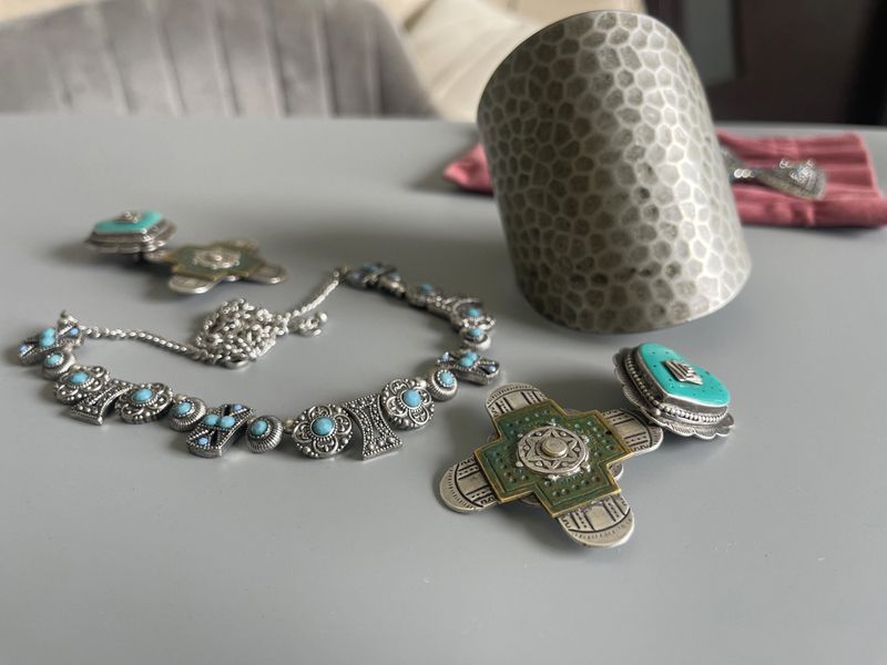 Various jewelry pieces are arranged on a table. There is a silver bracelet, a necklace, and an ornate cup made of metal. The setting is indoors with natural light.