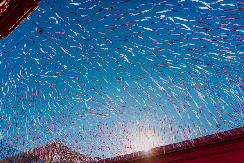Vibrant multicolored streamers stretch across a bright blue Oaxaca sky above colonial rooftops, sunlight peeking through; festive outdoor decorations capture the celebratory atmosphere of a Mexican street festival.
