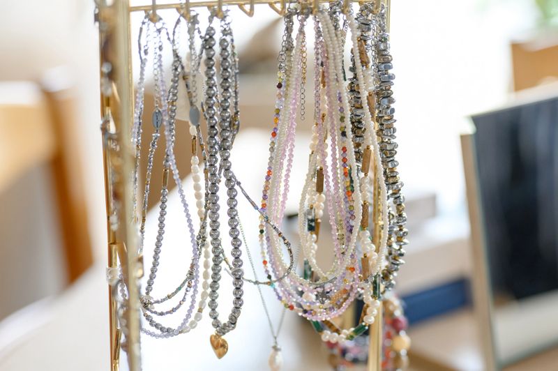 Various necklaces made of beads and pearls are displayed on a jewelry stand in a well-lit room with wooden furniture in the background