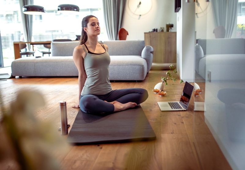 Mid adult Caucasian woman practicing yoga at home on a mat, using a laptop for guidance. She is seated with a calm expression, surrounded by a serene living space with natural lighting.