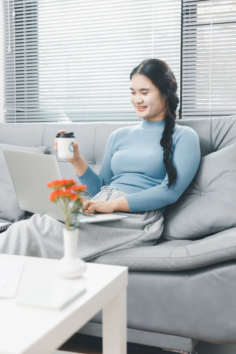 A smiling Asian woman sitting on a sofa with her laptop, holding a supplement bottle. She represents health-conscious remote workers taking care of their wellbeing while working at home.