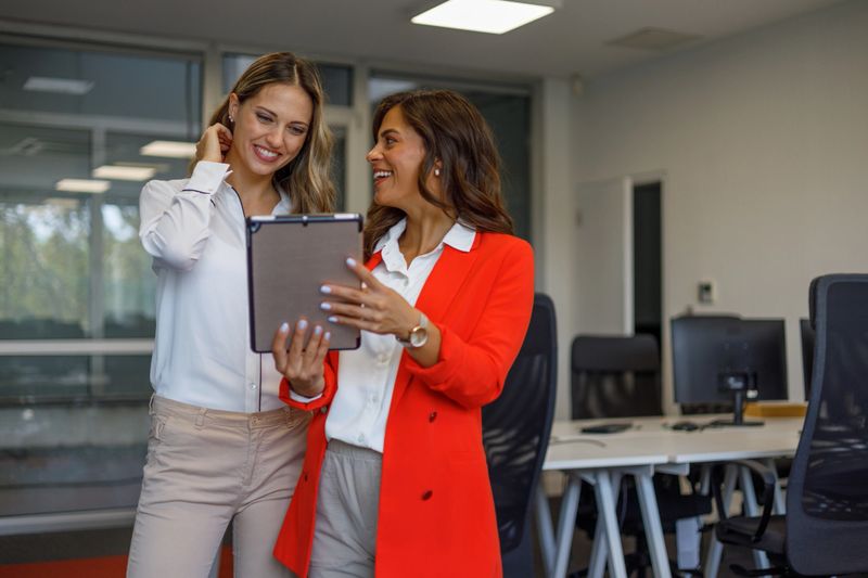 Two smiling adult businesswomen collaborating, discussing data on a digital tablet in a bright, contemporary office environment