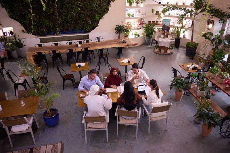 A multicultural group of coworkers meet casually in a plant-filled Dubai coffee shop, sharing laptops and notes while brainstorming, collaborating and enjoying coffee during a relaxed professional meeting.