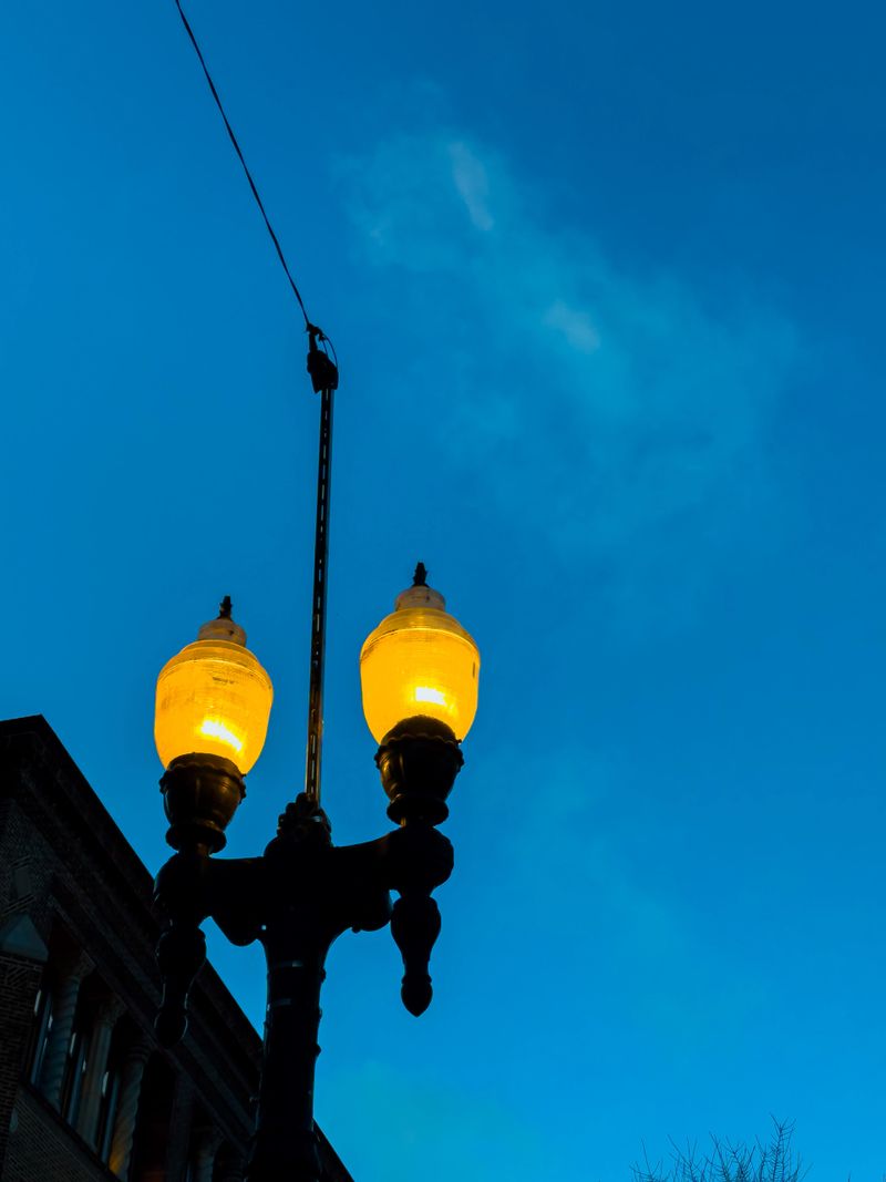A street lamp lights up as dusk falls in a city. The sky appears blue with clouds. The lamp is close to a building.