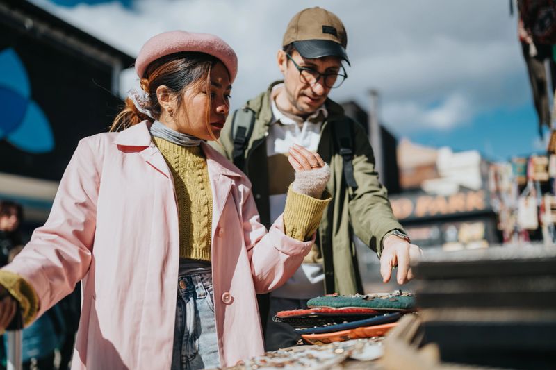 A multiethnic couple of tourists examines items at an outdoor market stall. The pair shops together while browsing handmade souvenirs and accessories.