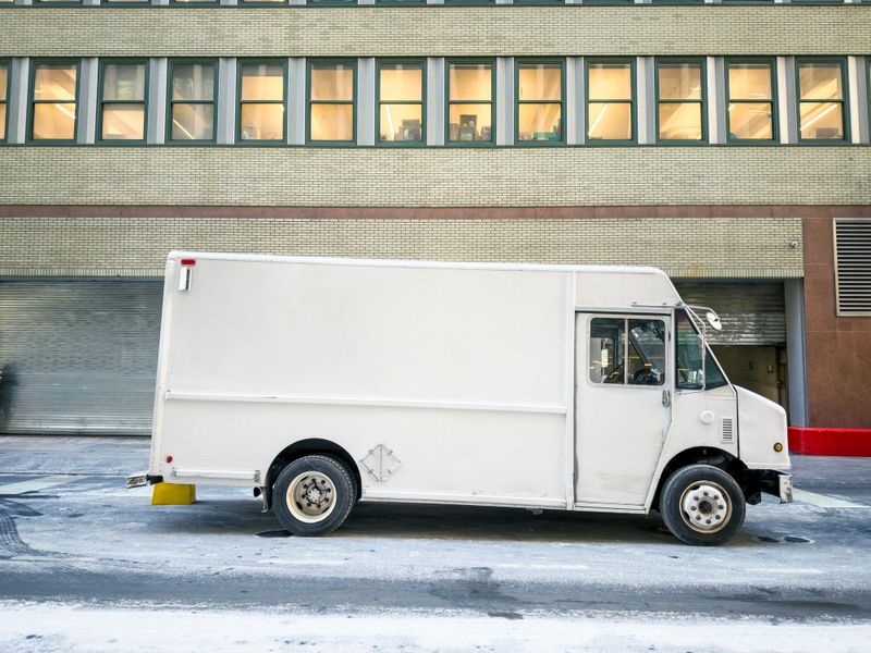 Full frame side view of plain white box delivery van parked curbside on light snow covered sidewalk beside beige brick commercial building with large windows during winter daylight offering realistic copy space. Urban street scene with truck rear door ramp and wheel details captures logistics in cold weather ideal for delivery service shipping or city commerce concepts. Horizontal composition highlights everyday transport for commercial visuals.