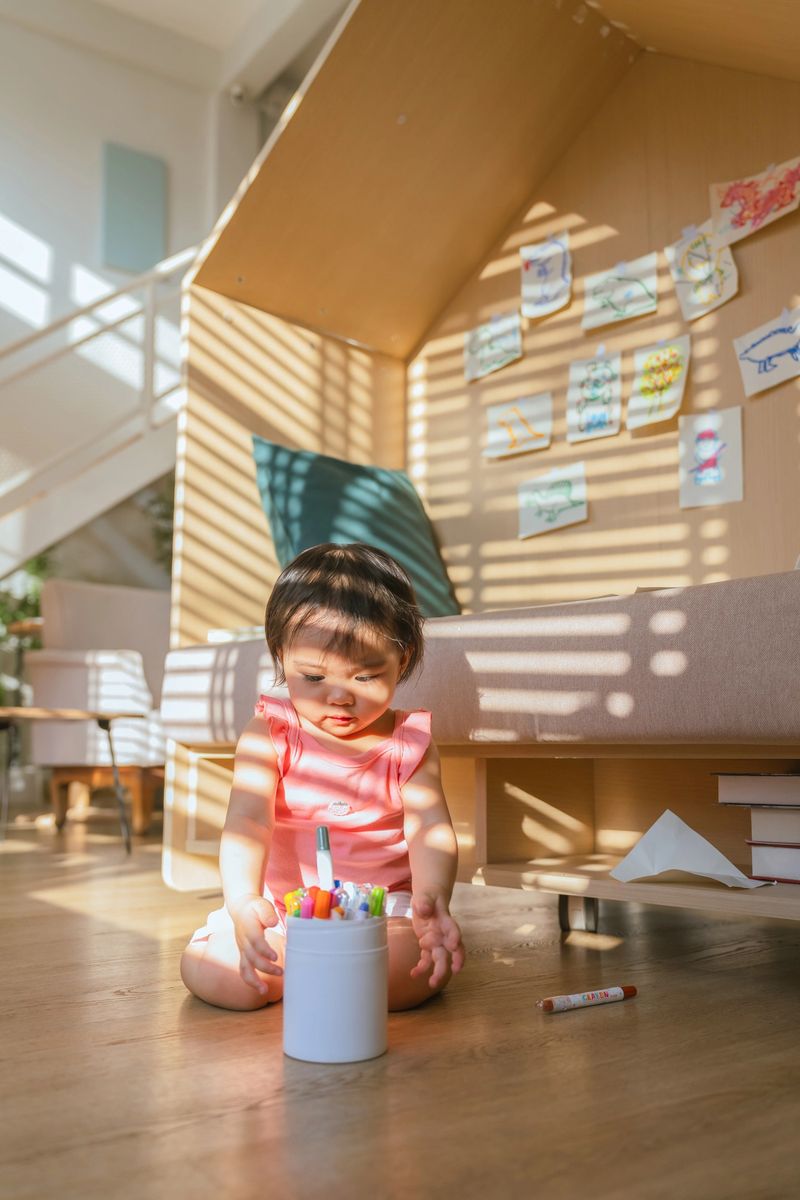 cute young Asian toddler girl in a pink romper sits on a sunlit wooden floor in a modern home, focused on creative drawing with colorful markers, capturing a moment of early childhood development