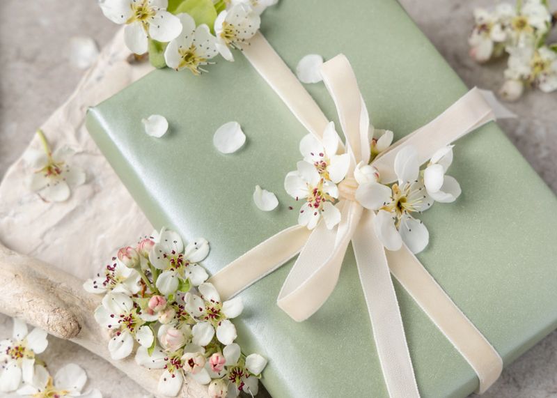 Light green wrapped gift box with white spring flowers and ribbon bow in soft pastel tones closeup. Pastel romantic present for birthday, wedding or seasonal celebration