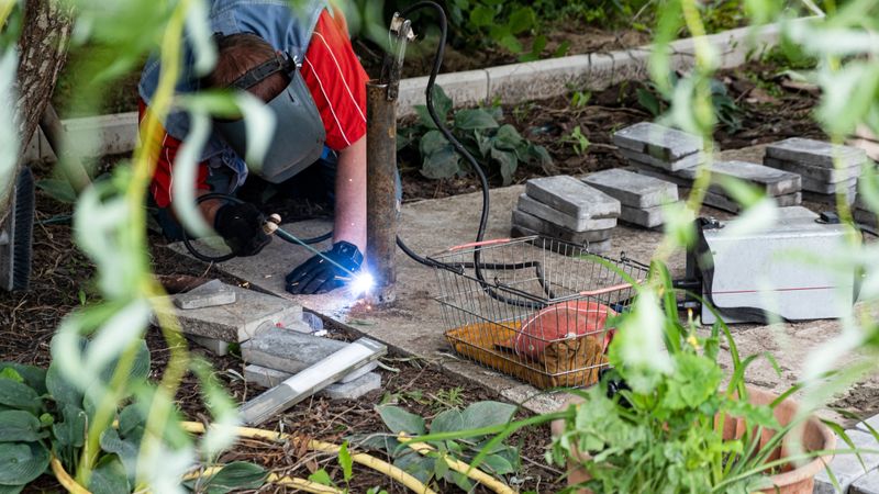 A male welder in full protective gear, including a welding helmet and gloves, is welding a metal foundation frame for a terrace next to a country house. The work takes place outdoors in a garden setting, with flowers and green plants visible nearby. Bright sparks fly from the welding point, contrasting with the peaceful natural background. The image combines themes of home construction, landscaping, and professional craftsmanship. Ideal for content on home improvement, outdoor building projects, terrace installation, garden renovation, and skilled manual labor.