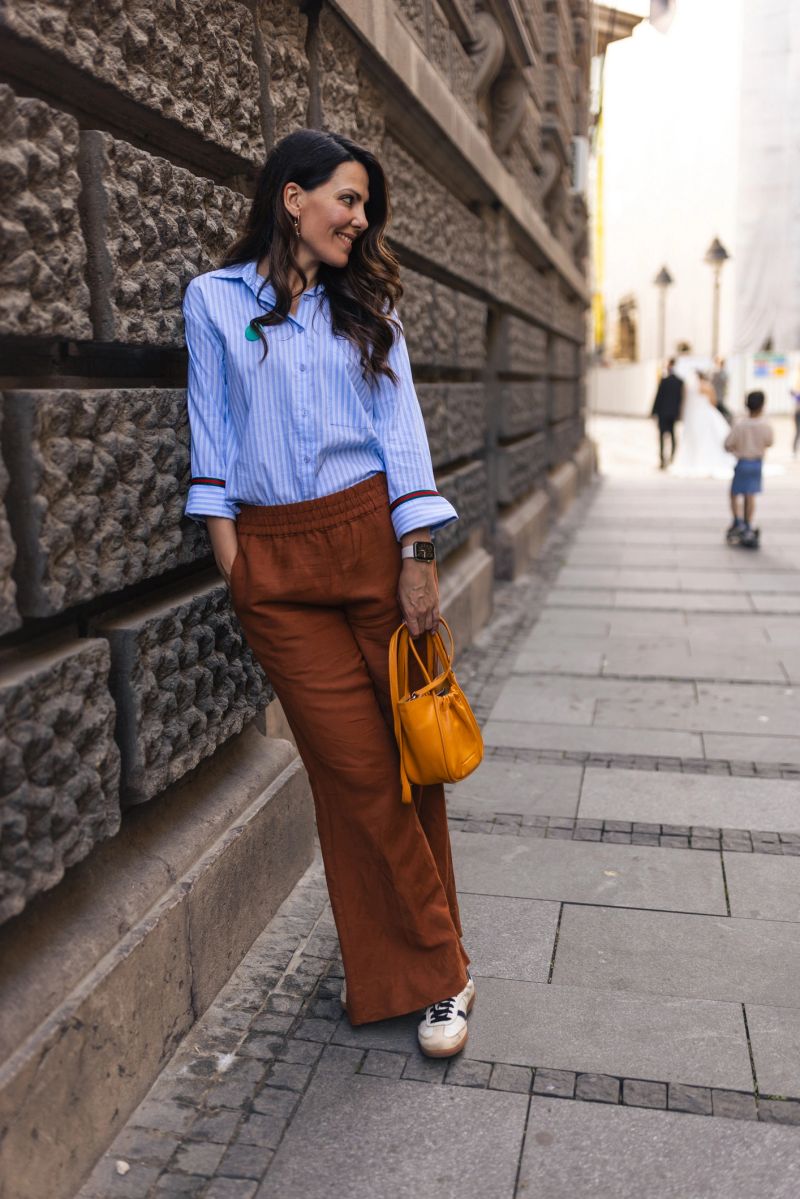 A fashionable woman in a blue shirt and brown trousers is walking down a historic street with a textured wall and cobblestone pathway, carrying a yellow handbag.