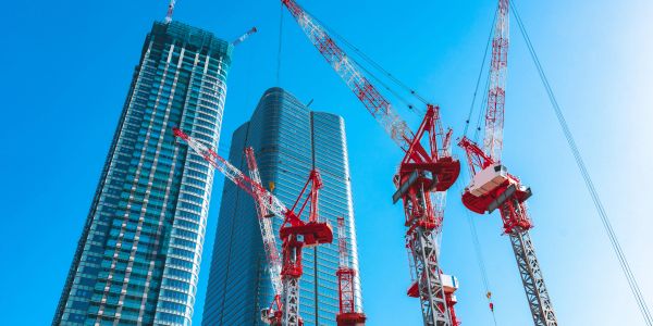 Construction cranes with tall skyscrapers under a clear blue sky.