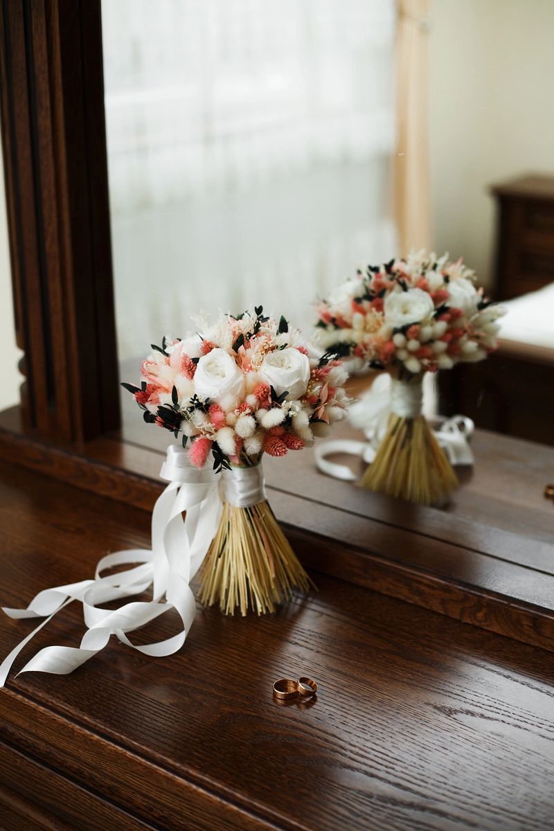A bouquet of flowers is placed on a wooden table. A mirror reflects the bouquet and a wedding ring rests next to it. The setting is bright and inviting with soft lighting.