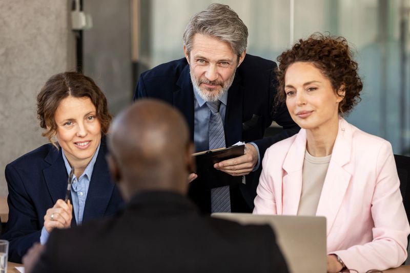 A diverse panel of professionals evaluates a candidate in a modern office setting, conveying a focused, collaborative business meeting and recruitment process with attentive, professional expressions.