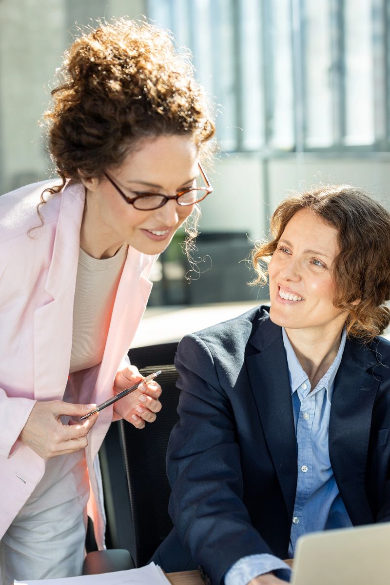 Two professional women collaborate over a laptop in a bright modern office, sharing ideas and smiling as they review documents and discuss work during a friendly business meeting.