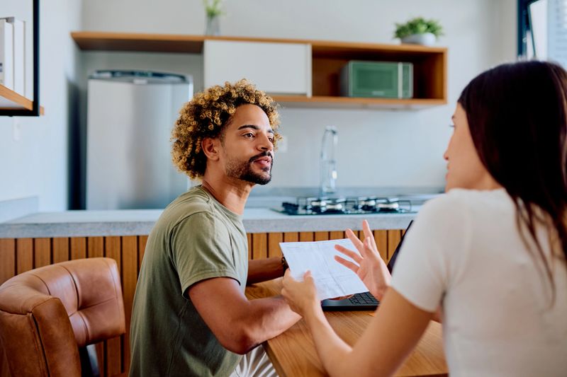 Young adult couple exchanging information and discussing their financial situation while going over documents and using a laptop in the kitchen