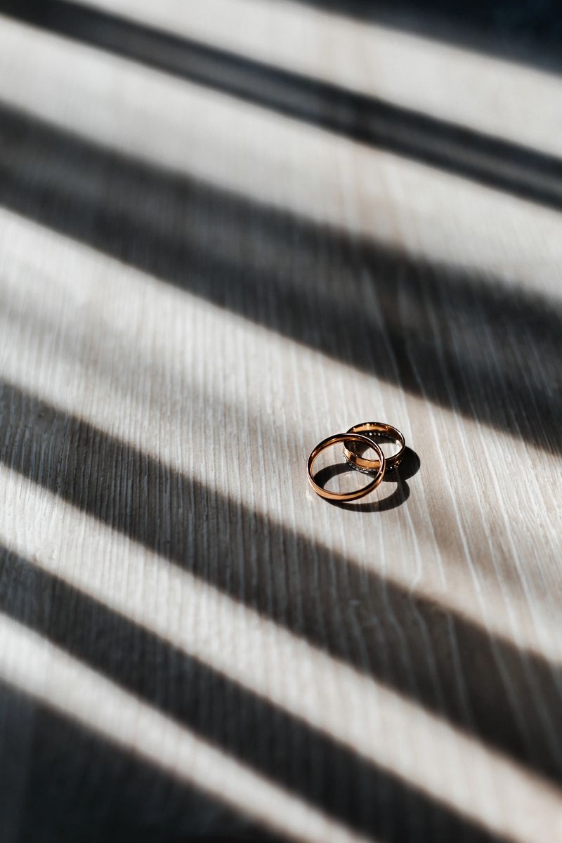 Two gold wedding rings rest on a wooden table. Light falls through the window, creating shadow lines on the surface. This scene is captured during the day.