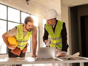 Two construction workers reviewing plans on a laptop.