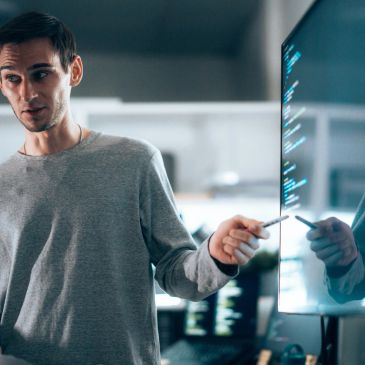 Man in gray sweater pointing at code on a transparent screen.