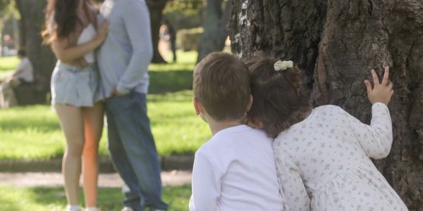 Two children hiding behind a tree, watching a couple in a park.