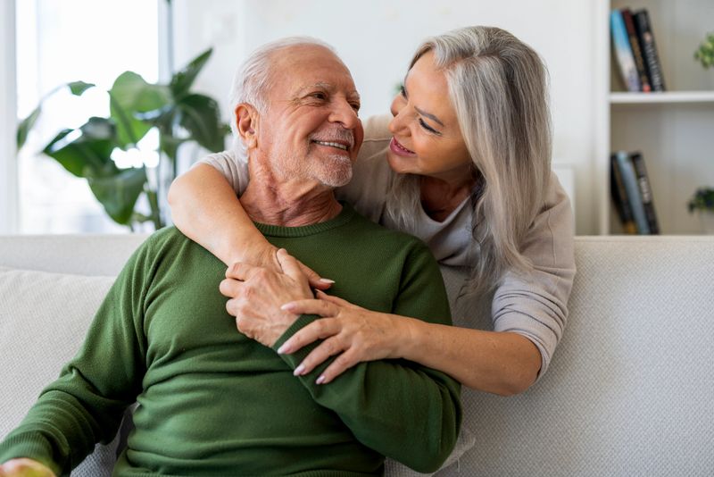 Senior couple smiling and hugging on a sofa at home, enjoying a warm and intimate moment in a cozy living room.