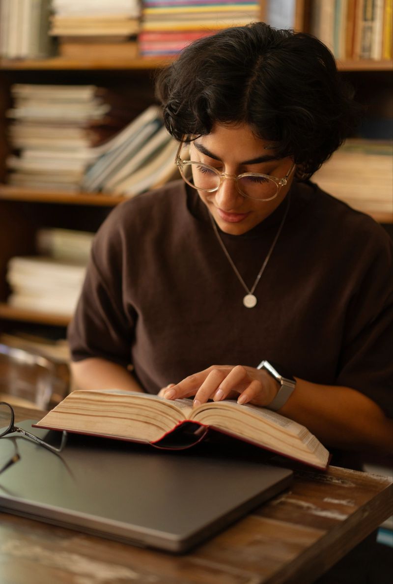 A young woman reading a book at her desk in a warm library environment.