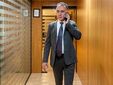 Man in a suit talking on a phone in an office corridor.