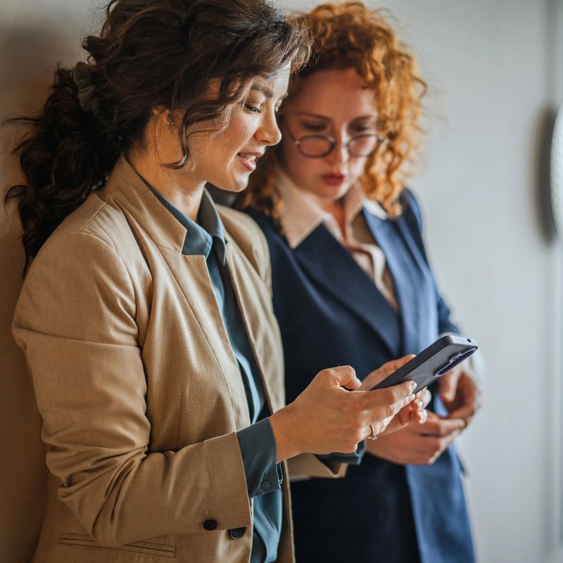Two professional businesswomen collaborating and engaging in a casual discussion while looking at a smart phone screen, representing modern communication and teamwork in an office setting