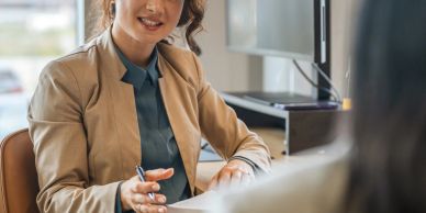 Professional woman in a beige blazer discussing with a colleague at office desk.