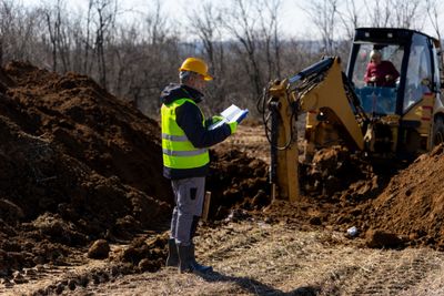 Construction worker inspecting site with excavator in background.