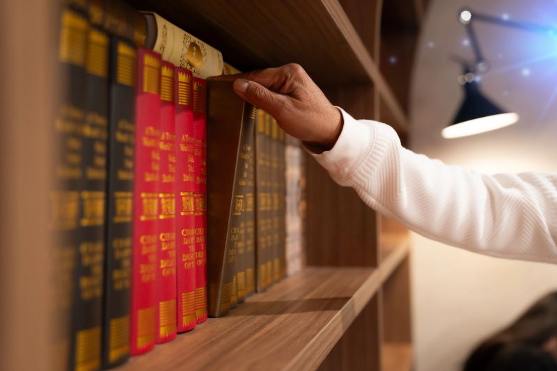 Close-up of a hand pulling a brown hardcover book from a wooden bookshelf filled with colorful books.