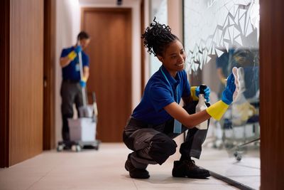 Two professional cleaners cleaning glass and floors in an office corridor.
