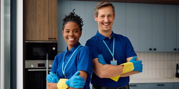 Two cheerful cleaners in blue uniforms with rubber gloves stand in a kitchen.