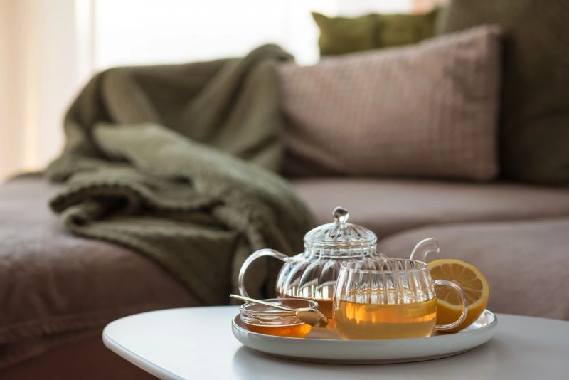 Glass teapot with amber tea, teacup with lemon slice, and bowl of honey with golden spoon on white table in cozy living room