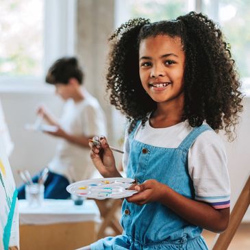 Girl holding paint palette and brush smiling at camera.
