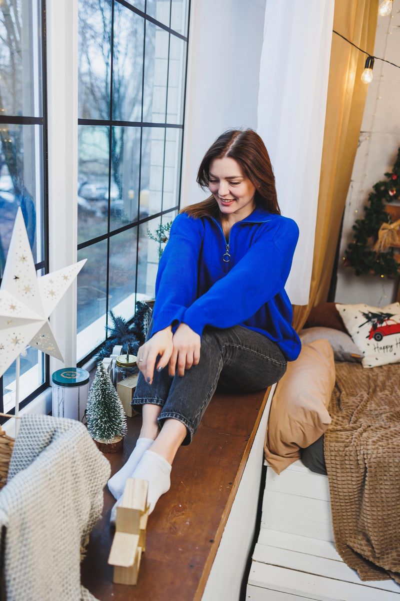 A young pretty woman in a blue knitted sweater is sitting by the window waiting for the new year. New Year's mood in a home atmosphere.
