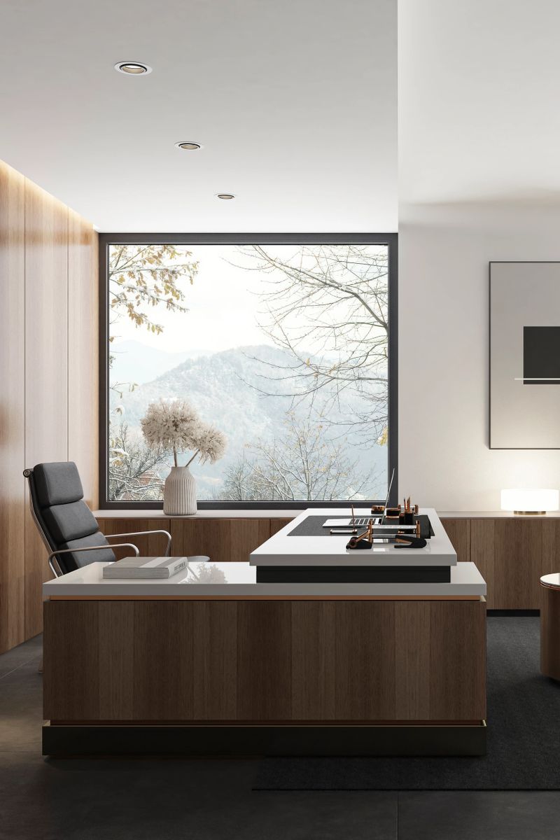 Modern luxury executive office with warm oak walnut wood-paneled walls, built-in dark shelving, and a large wood and white L-shaped desk with a laptop and black leather chair. A large window reveals a snowy mountain landscape. Two dark grey guest chairs sit near a cylindrical wooden side table. A low credenza holds a glowing lamp and dried floral vases beneath minimalist abstract art. Soft natural daylight and warm recessed lighting, elegant professional atmosphere. Modern and luxury Work place.