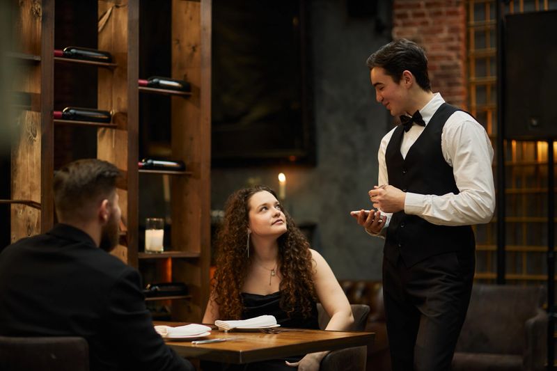 Young adult waiter taking order from Caucasian couple in restaurant, using handheld device for service. Ideal for hospitality marketing, dining service, customer experience campaigns