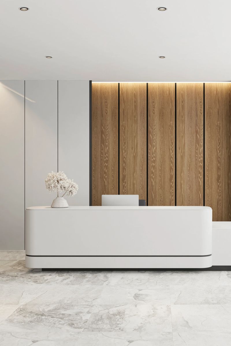 Contemporary reception area with a sleek white curved desk set against a warm vertical wooden slat feature wall with integrated ambient lighting. Surrounding walls are finished in smooth, glossy light-toned panels, complemented by a textured stone accent wall on the side. The ceiling features a recessed geometric design with built-in spotlights, enhancing the clean architectural lines. The floor is covered with large-format white gray stone tiles, creating a refined contrast with the light interior palette. Styled with minimal decor and neutral tones, the space conveys a calm, professional, and luxurious atmosphere. Ideal for use in interior design presentations, office and corporate branding, architectural visualization, real estate marketing, hospitality projects, and modern workspace concepts.