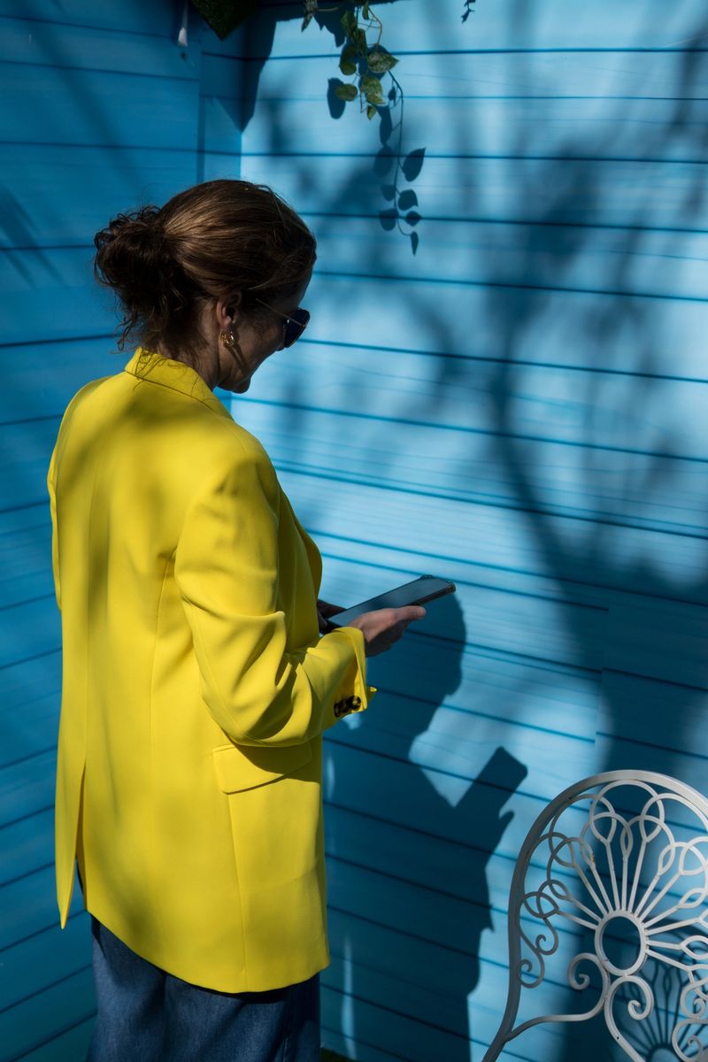 Back view of a woman dressed in a bright yellow blazer standing in front of a light blue wooden wall decorated with green artificial leaves and shadows.