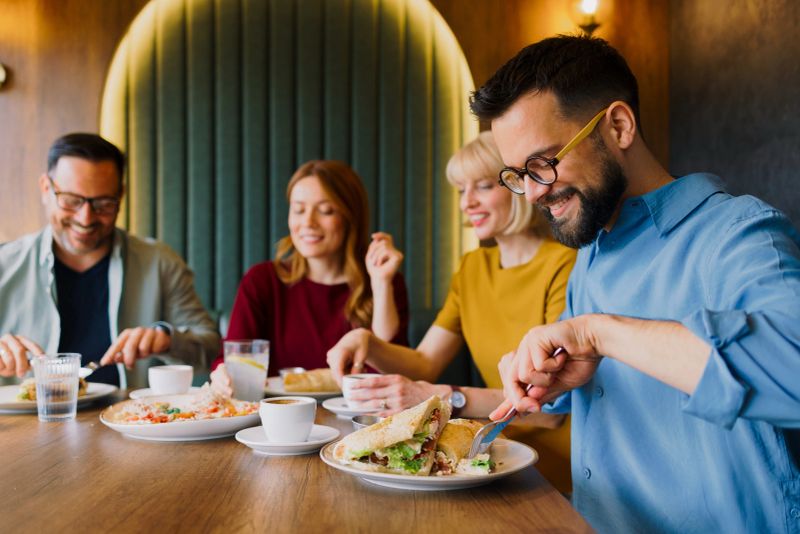 Group of young adults enjoying meal together, eating sandwiches and drinking coffee in cozy restaurant.